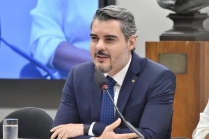 Homem de terno e gravata falando ao microfone durante uma reunião ou audiência pública, sentado à mesa, com expressão séria e gesticulando com as mãos. Ao fundo, há uma tela e elementos de um ambiente institucional.