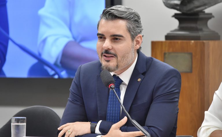 Homem de terno e gravata falando ao microfone durante uma reunião ou audiência pública, sentado à mesa, com expressão séria e gesticulando com as mãos. Ao fundo, há uma tela e elementos de um ambiente institucional.