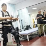 Pessoa com lesões na medula espinhal utilizando um exoesqueleto robótico para reabilitação motora, em ambiente clínico, enquanto profissionais e jornalistas ao redor observam e registram a demonstração com câmeras.