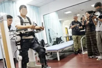 Pessoa com lesões na medula espinhal utilizando um exoesqueleto robótico para reabilitação motora, em ambiente clínico, enquanto profissionais e jornalistas ao redor observam e registram a demonstração com câmeras.