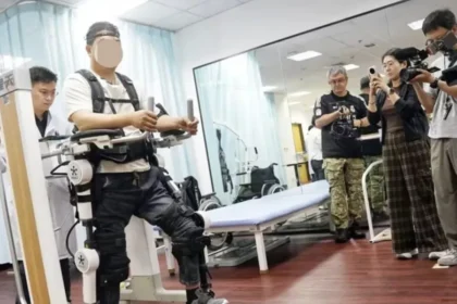 Pessoa com lesões na medula espinhal utilizando um exoesqueleto robótico para reabilitação motora, em ambiente clínico, enquanto profissionais e jornalistas ao redor observam e registram a demonstração com câmeras.