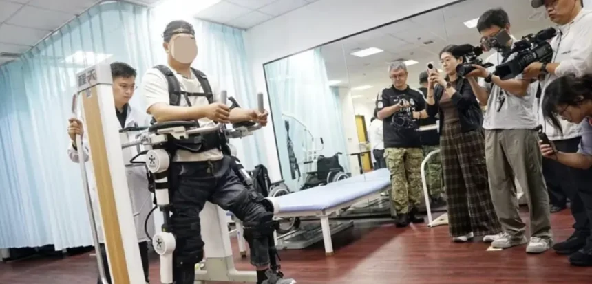 Pessoa com lesões na medula espinhal utilizando um exoesqueleto robótico para reabilitação motora, em ambiente clínico, enquanto profissionais e jornalistas ao redor observam e registram a demonstração com câmeras.