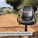 Imagem mostra uma cadeira de escritório preta com a palavra “Previdência” escrita no encosto, posicionada no meio de uma rua de terra em São Sebastião. Ao fundo aparecem casas, árvores e áreas sem pavimentação. Na parte inferior da imagem, uma legenda informa que a rua é a única da região sem asfalto.