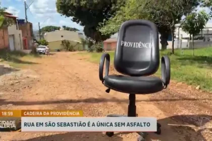 Imagem mostra uma cadeira de escritório preta com a palavra “Previdência” escrita no encosto, posicionada no meio de uma rua de terra em São Sebastião. Ao fundo aparecem casas, árvores e áreas sem pavimentação. Na parte inferior da imagem, uma legenda informa que a rua é a única da região sem asfalto.