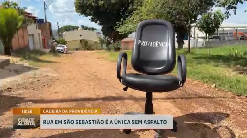 Imagem mostra uma cadeira de escritório preta com a palavra “Previdência” escrita no encosto, posicionada no meio de uma rua de terra em São Sebastião. Ao fundo aparecem casas, árvores e áreas sem pavimentação. Na parte inferior da imagem, uma legenda informa que a rua é a única da região sem asfalto.