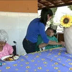 **Texto alternativo (breve):** Cuidadora auxilia uma pessoa idosa durante uma refeição, enquanto outra idosa sentada à mesa se alimenta. O ambiente é simples e decorado com flores.