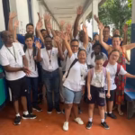 Grupo de estudantes com deficiência e educadores posa em corredor escolar, sorrindo e acenando para a câmera.