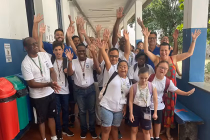 Grupo de estudantes com deficiência e educadores posa em corredor escolar, sorrindo e acenando para a câmera.
