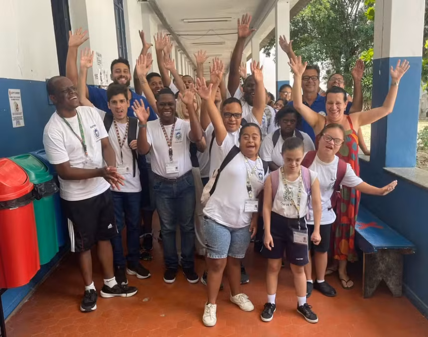 Grupo de estudantes com deficiência e educadores posa em corredor escolar, sorrindo e acenando para a câmera.