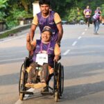 Pessoa em cadeira de rodas participa de uma corrida de rua com o apoio de um acompanhante, enquanto outros corredores seguem ao fundo.