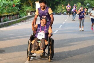 Pessoa em cadeira de rodas participa de uma corrida de rua com o apoio de um acompanhante, enquanto outros corredores seguem ao fundo.