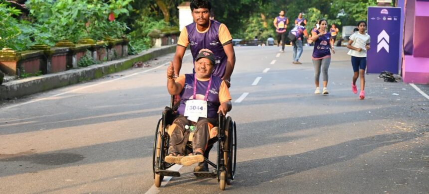 Pessoa em cadeira de rodas participa de uma corrida de rua com o apoio de um acompanhante, enquanto outros corredores seguem ao fundo.