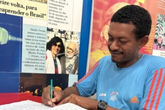 Homem escreve em um livro em braile sobre uma mesa. Ao fundo, há um mural com fotografias e referências ao educador Paulo Freire, destacando a importância da alfabetização, da educação inclusiva e do acesso à leitura para pessoas com deficiência visual.