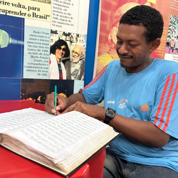Homem escreve em um livro em braile sobre uma mesa. Ao fundo, há um mural com fotografias e referências ao educador Paulo Freire, destacando a importância da alfabetização, da educação inclusiva e do acesso à leitura para pessoas com deficiência visual.