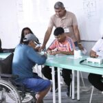 Eleitores com Deficiência: Pessoa em cadeira de rodas participa do atendimento durante um mutirão de cadastramento eleitoral. Ao redor, servidores e eleitores recebem orientações em uma sala de atendimento