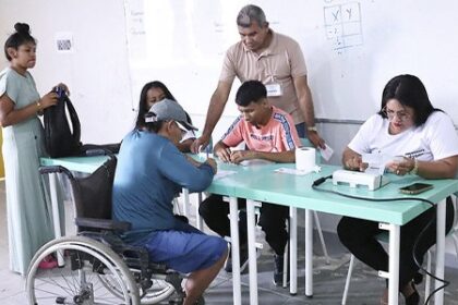 Eleitores com Deficiência: Pessoa em cadeira de rodas participa do atendimento durante um mutirão de cadastramento eleitoral. Ao redor, servidores e eleitores recebem orientações em uma sala de atendimento