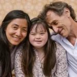 Um close-up de uma família interracial, incluindo uma mulher asiática, uma jovem com síndrome de Down e um homem branco. Eles estão sentados próximos e sorrindo. O homem tem o braço em volta da jovem e está encostado nela. A mulher e a jovem estão olhando para a câmera.