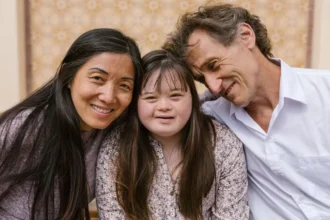 Um close-up de uma família interracial, incluindo uma mulher asiática, uma jovem com síndrome de Down e um homem branco. Eles estão sentados próximos e sorrindo. O homem tem o braço em volta da jovem e está encostado nela. A mulher e a jovem estão olhando para a câmera.