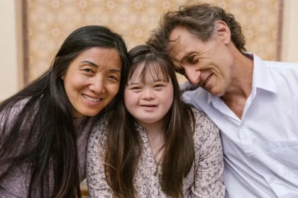 Um close-up de uma família interracial, incluindo uma mulher asiática, uma jovem com síndrome de Down e um homem branco. Eles estão sentados próximos e sorrindo. O homem tem o braço em volta da jovem e está encostado nela. A mulher e a jovem estão olhando para a câmera.
