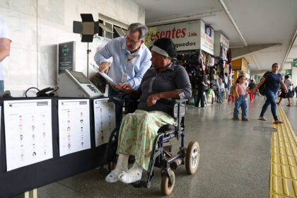 Um funcionário da Justiça Eleitoral orienta uma mulher cadeirante com bandagens nos pés na urna eletrônica.
