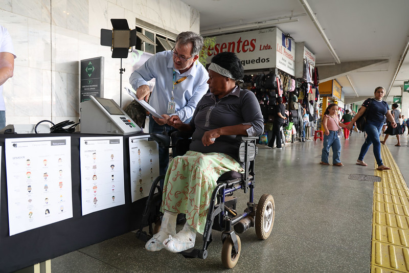 Um funcionário da Justiça Eleitoral orienta uma mulher cadeirante com bandagens nos pés na urna eletrônica.