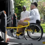 **Texto Alt:** Homem em cadeira de rodas utilizando o elevador hidráulico de uma van acessível, acompanhado por um motorista ao fundo.