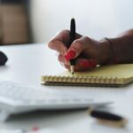 Mão com unhas vermelhas escreve em bloco amarelo sobre mesa branca, próxima a teclado e mouse.