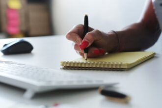 Mão com unhas vermelhas escreve em bloco amarelo sobre mesa branca, próxima a teclado e mouse.