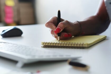 Mão com unhas vermelhas escreve em bloco amarelo sobre mesa branca, próxima a teclado e mouse.