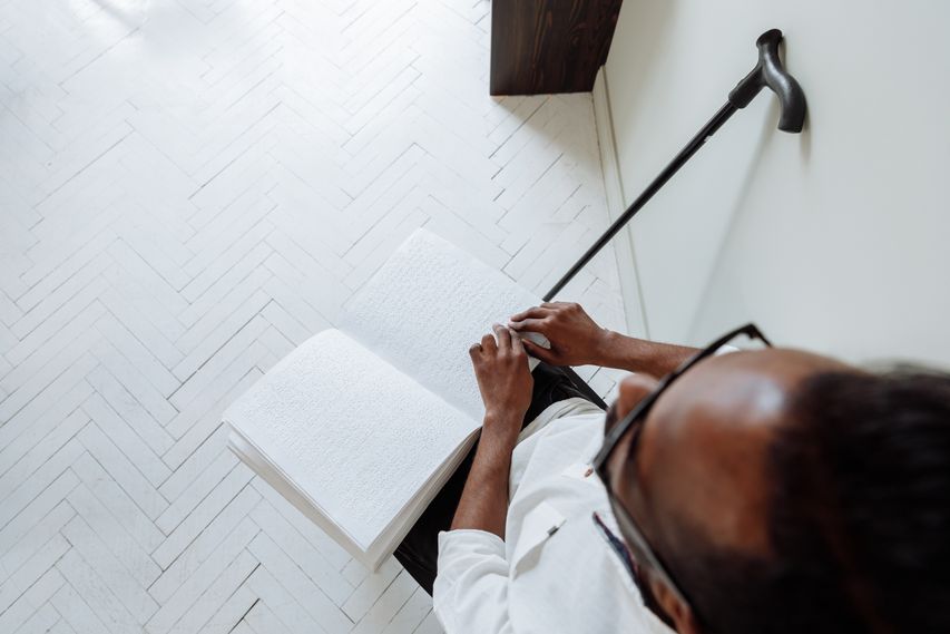Vista superior de um homem de óculos lendo um livro em Braille, com uma bengala ao lado.