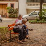 Casal de idosos aproveitando um momento tranquilo em um banco de parque em um ambiente urbano com carros e vegetação