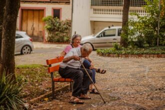 Casal de idosos aproveitando um momento tranquilo em um banco de parque em um ambiente urbano com carros e vegetação