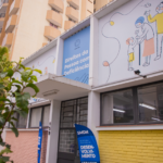 Fachada do Departamento dos Direitos da Pessoa com Deficiência, em Curitiba (PR), com painel azul de identificação, ilustrações de pessoas de diferentes idades, janelas de vidro e placas da Secretaria Municipal de Direitos Humanos (SMDH) na entrada.