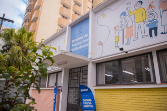 Fachada do Departamento dos Direitos da Pessoa com Deficiência, em Curitiba (PR), com painel azul de identificação, ilustrações de pessoas de diferentes idades, janelas de vidro e placas da Secretaria Municipal de Direitos Humanos (SMDH) na entrada.