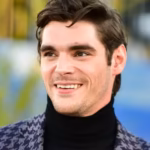 Ator RJ Mitte, pessoa com deficiência e conhecido por sua participação na série Breaking Bad, aparece sorridente, usando blusa preta de gola alta e blazer estampado, com cabelos castanhos penteados para o lado e fundo desfocado.