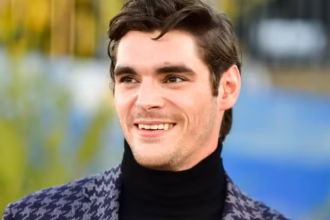 Ator RJ Mitte, pessoa com deficiência e conhecido por sua participação na série Breaking Bad, aparece sorridente, usando blusa preta de gola alta e blazer estampado, com cabelos castanhos penteados para o lado e fundo desfocado.