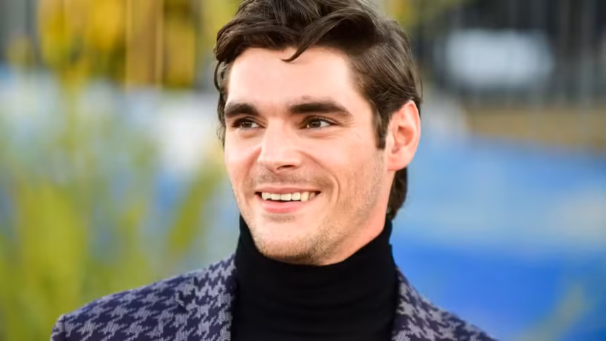 Ator RJ Mitte, pessoa com deficiência e conhecido por sua participação na série Breaking Bad, aparece sorridente, usando blusa preta de gola alta e blazer estampado, com cabelos castanhos penteados para o lado e fundo desfocado.
